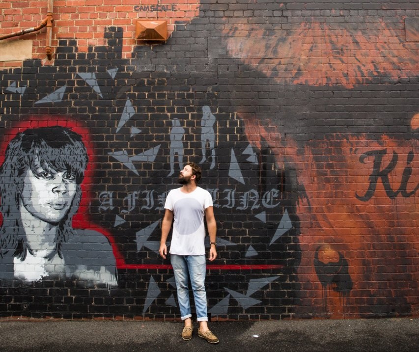 A man stands underneath a brick wall filled with street art. He wears a white t-shirt and light denim jeans.