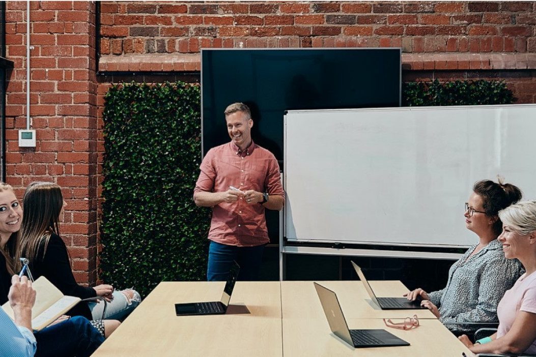 A person stands at the front of a meeting room in front of a whiteboard. A table full of people sit listening to them talk.