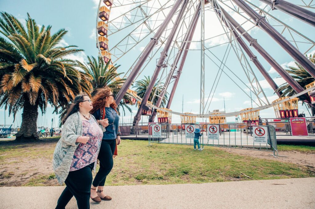 Two women walk together, both looking at a ferris wheel behind them.