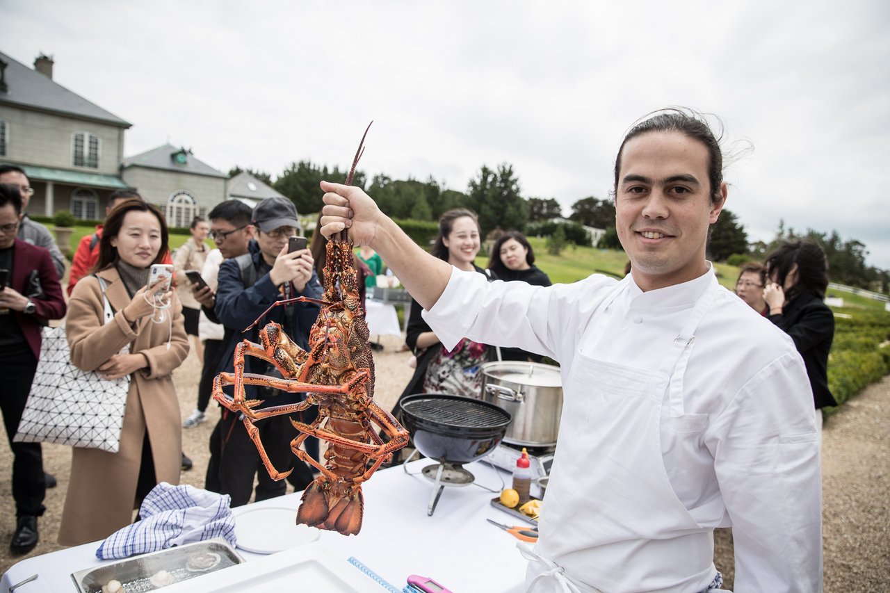 Chef holds a lobster in the air with a group of people looking on in background.