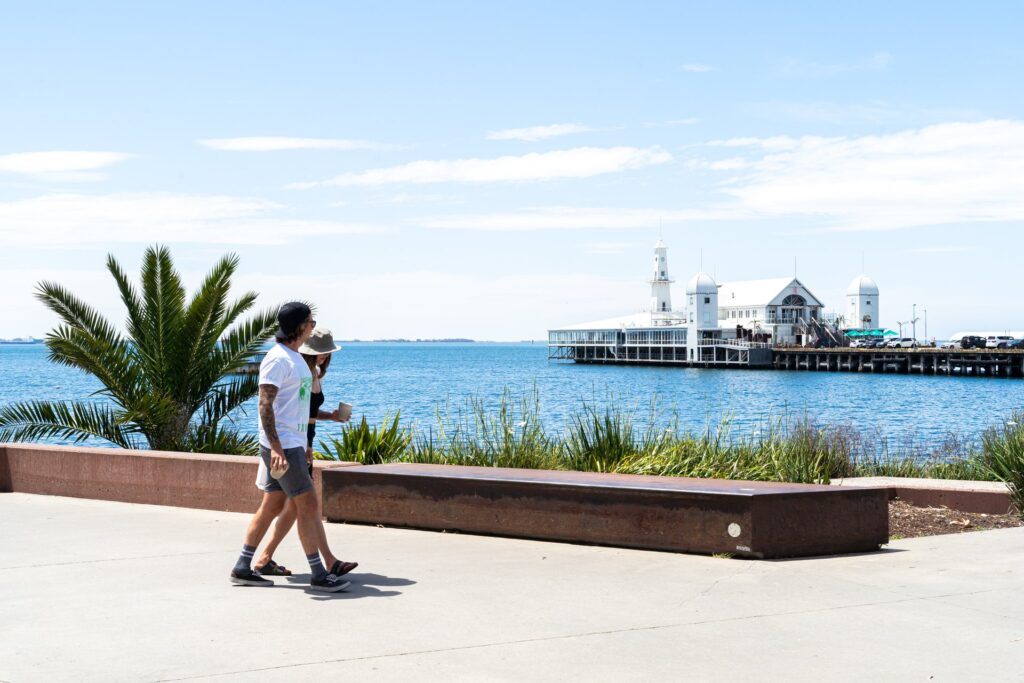 Two people walking along waterfront, a palm tree and pier are in the background.