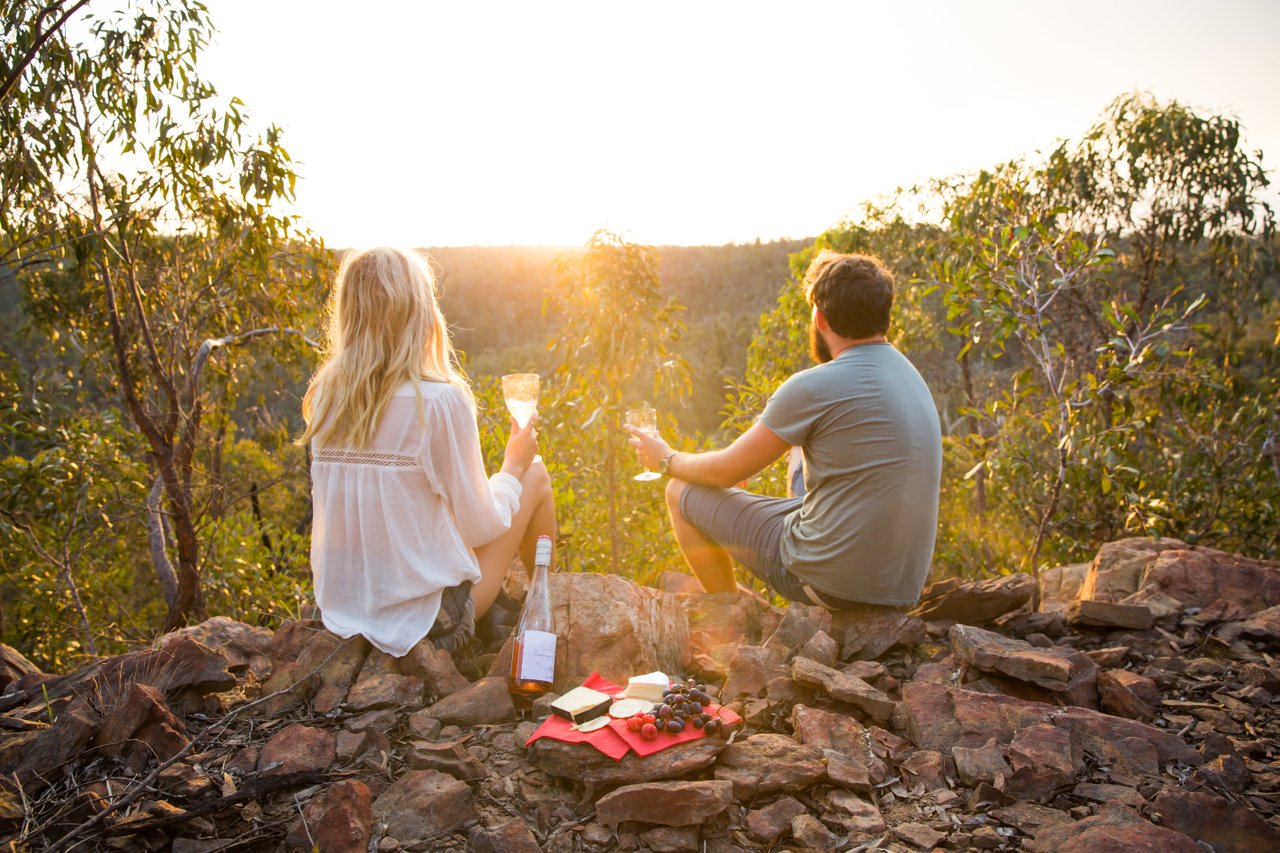 A man and woman sit drinking wine on the edge of a cliff, overlooking the sunset in the Brisbane Ranges.
