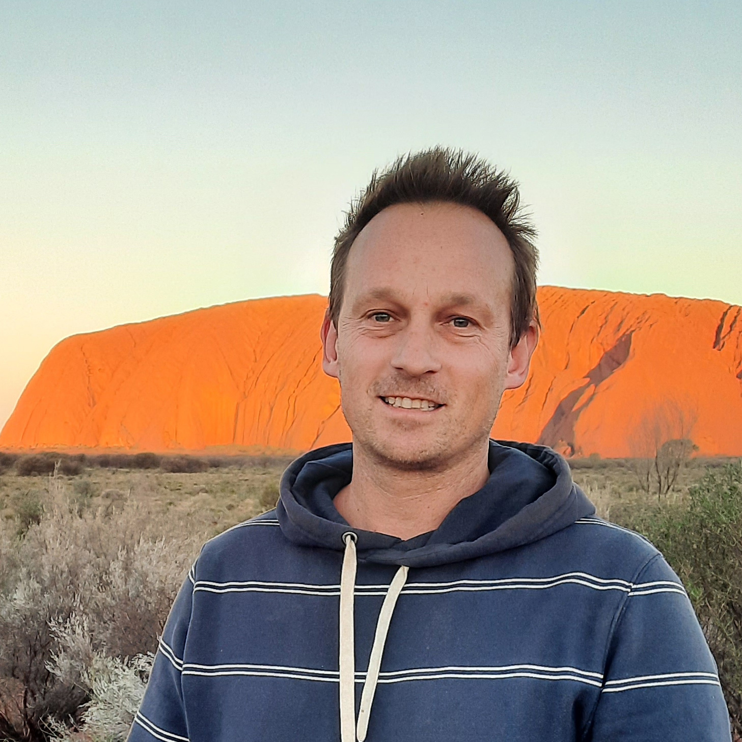 Tim stands in front of the uluru wearing a navy striped sweater. He has short brown hair. 