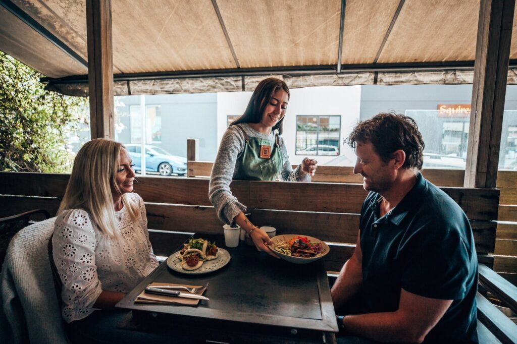 A couple sits at an outdoor table under a verandah at a cafe. A waitress is placing their meals on the table and the couple smiles up at her.