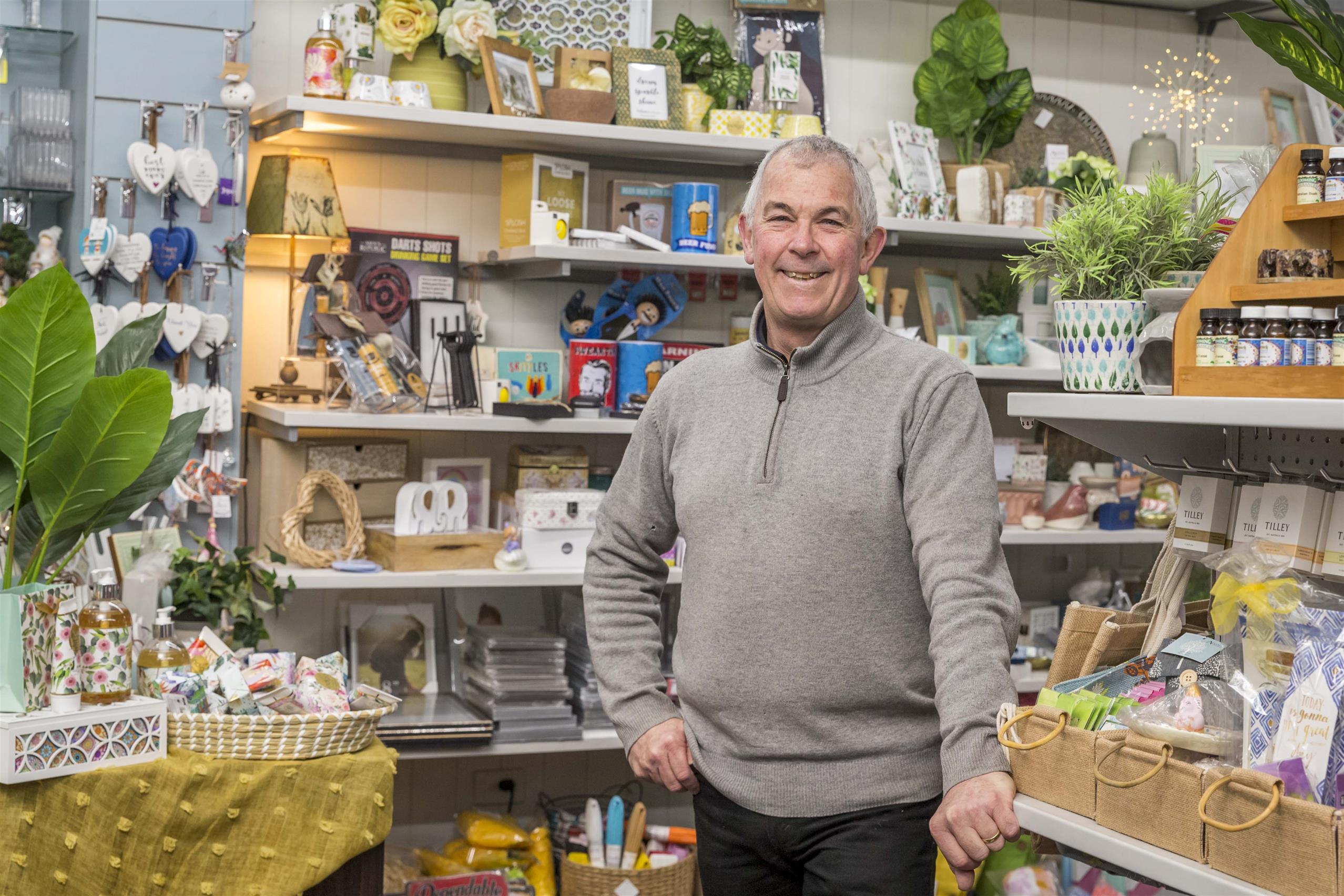 A man stands beside his shop counter smiling at the camera. Shelves full of giftware, nick nacks and decor fill the space behind him.
