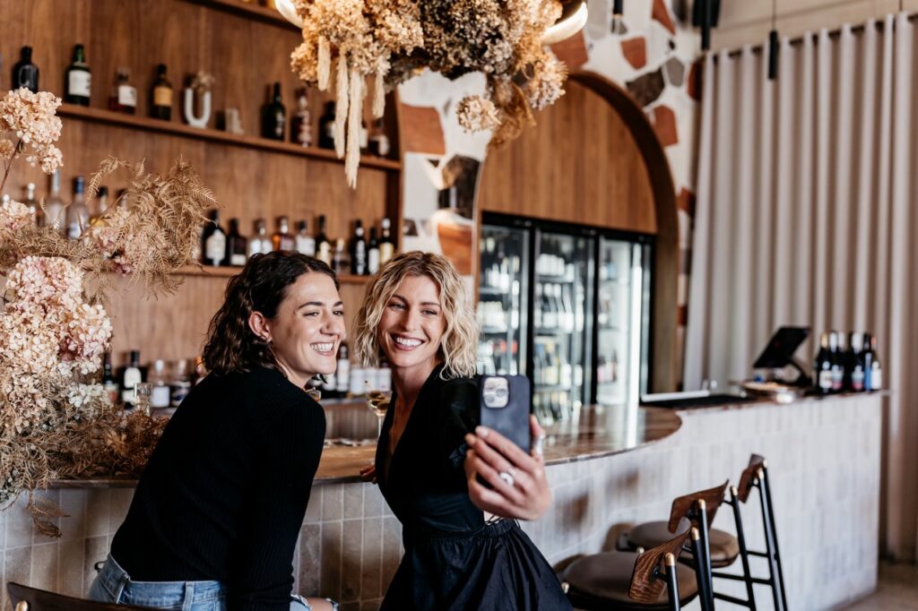 Two women post for a selfie while sitting at a bar.