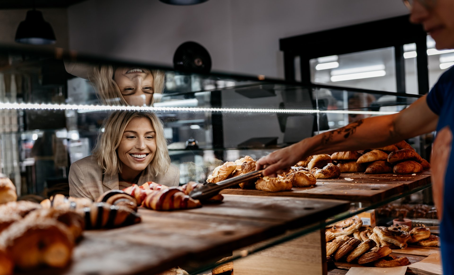 A woman with short blonde hair looks through a glass pastry cabinet as a worker picks up a croissant with tongs.