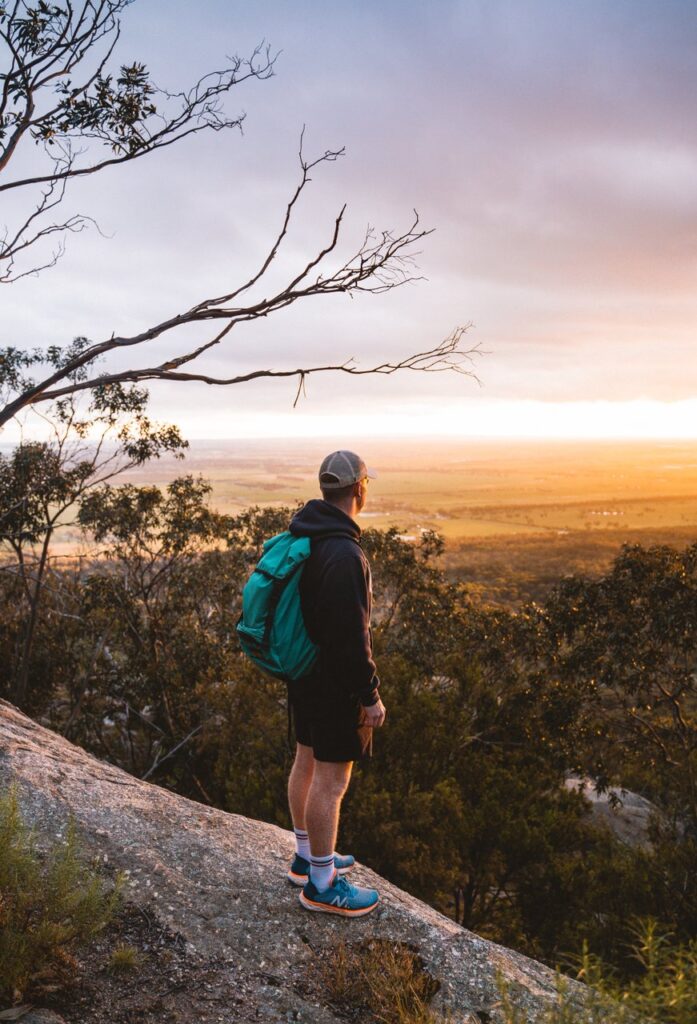 A man stands at the top of the You Yangs overlooking the view and sunset. He wears a cap, back pack and sports clothing.