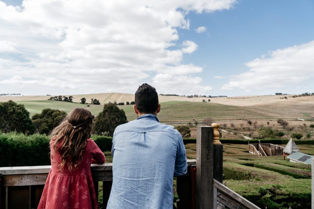 A man and young girl stand facing away from the camera. They look over a balcony towards rolling grassy hills and blue skies.