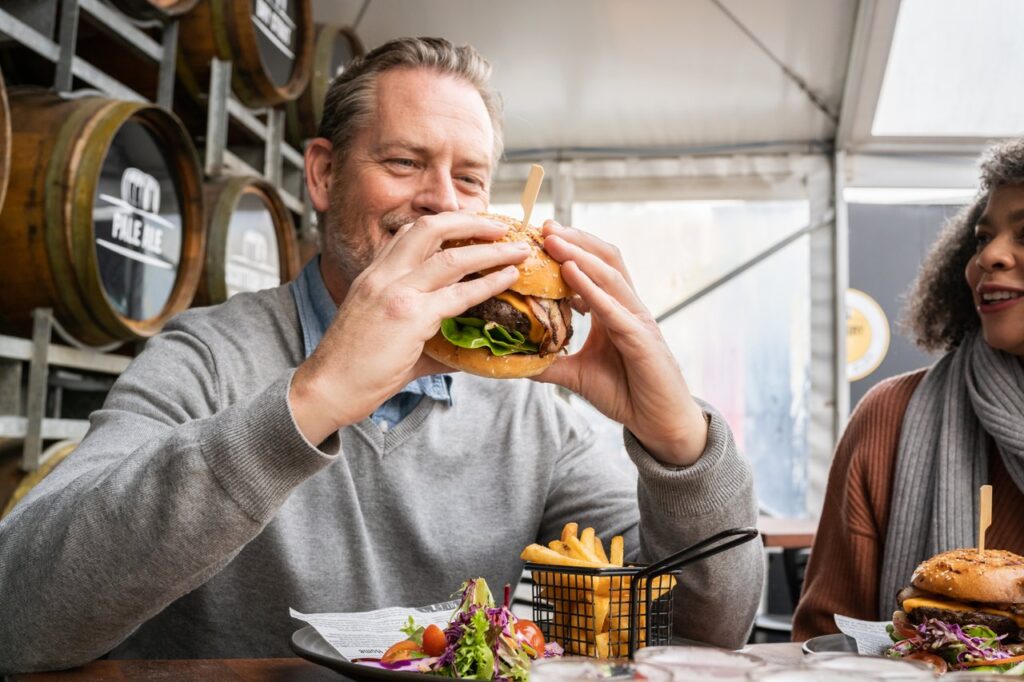 A man sits at a table holding a burger in both hands about to take a bit. A woman sits next to him watching with a smile.