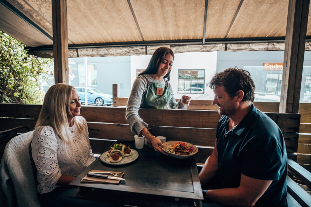 A cafe worker passes two plates of food to a man and woman sitting at a table.