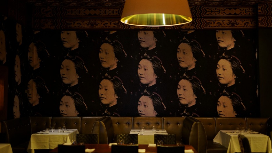 A dark leather booth is in the centre of the shot, with dark wallpaper featuring the face of a young woman repeated. A gold pendant light hangs just in shot with dim lighting.