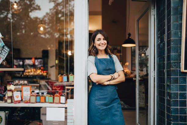 A business owner wears an apron and leans against the door frame at the front of her cafe.