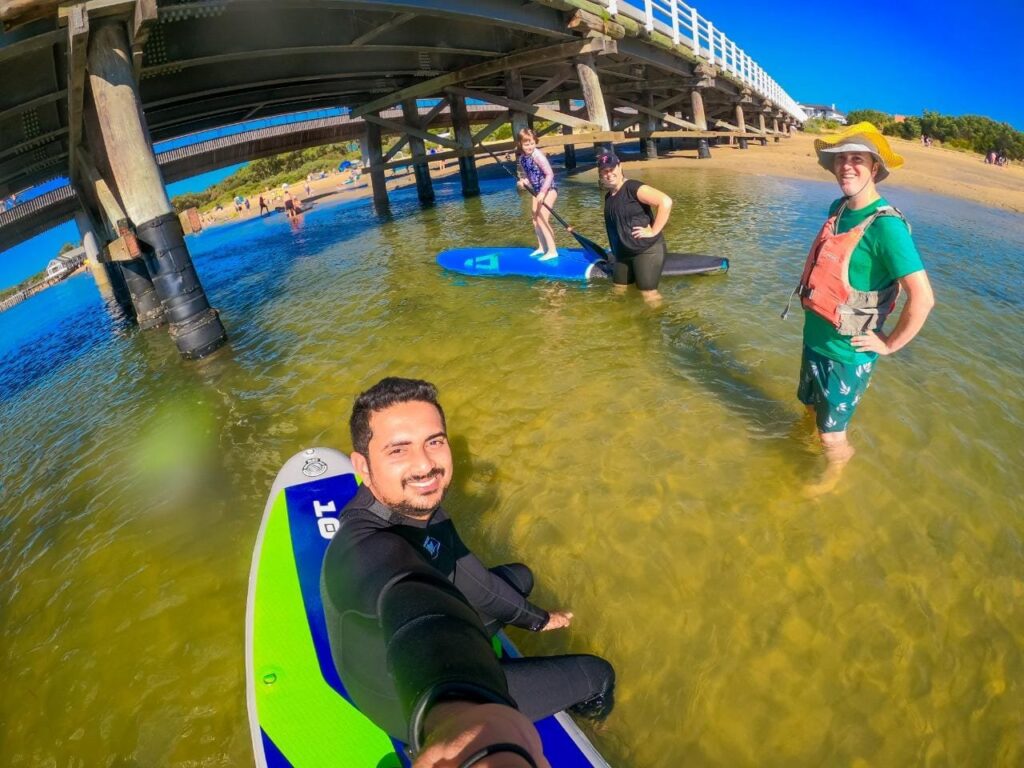 Four people stand in the shallows of a river posing for a photo, two people are on stand up paddle boards. A bridge crosses the river behind them.