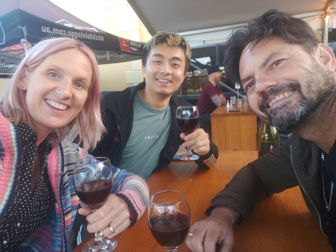 Three people sit on a table at a winery posing for a photo. They all hold glasses of red wine.