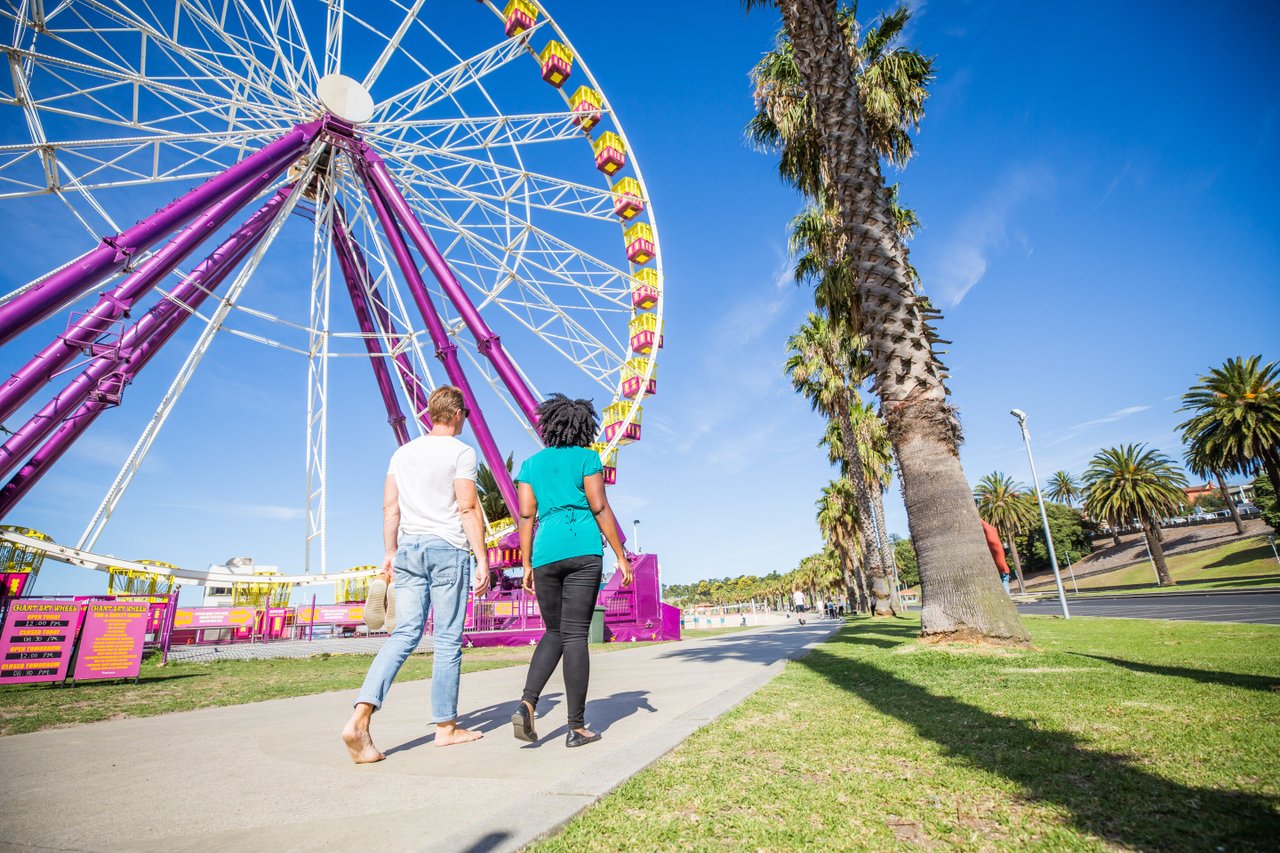A man and woman walk away from the camera down a path alongside a ferris wheel and large palm trees. The sky is blue and a glimpse of the bay is in the background.