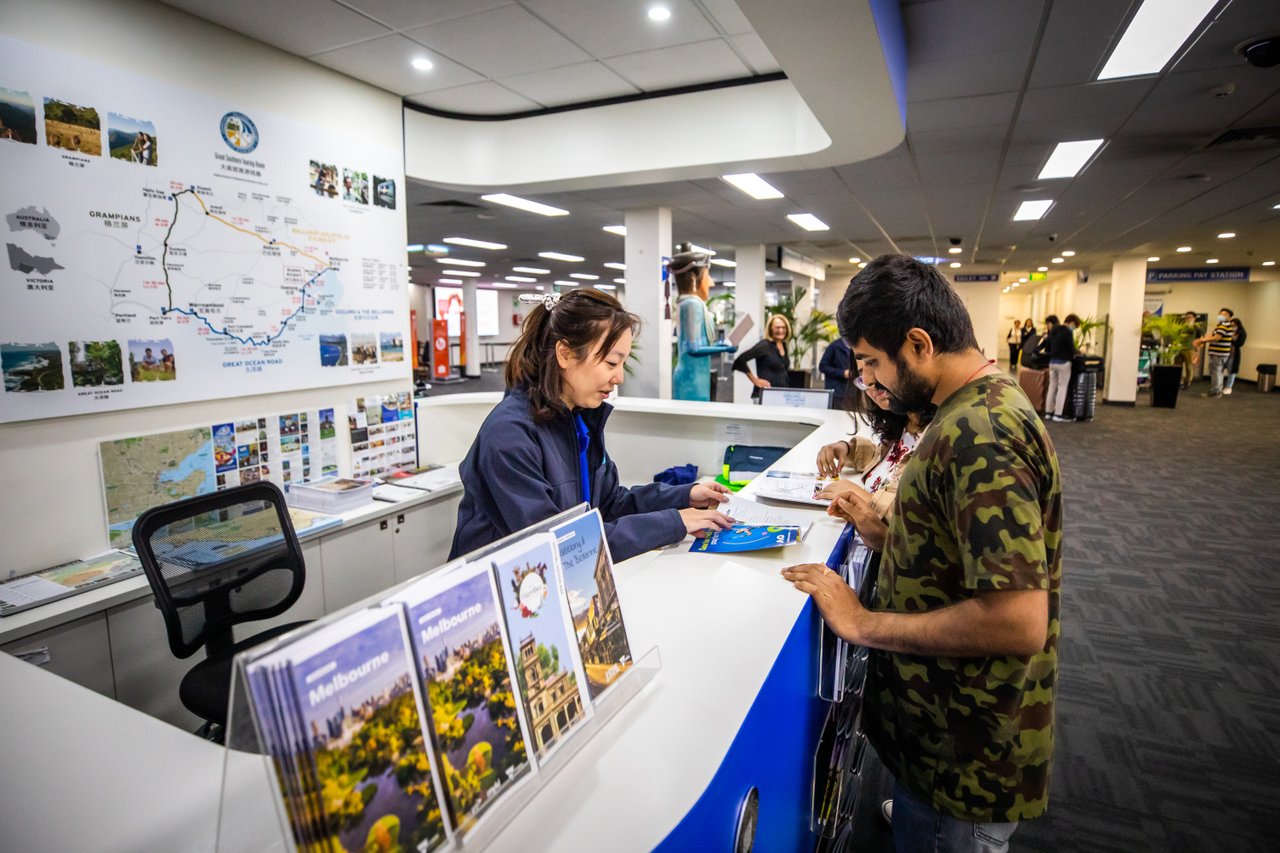 A woman speaks with a man at an information counter. They both look at a brochure that is on the counter between them.