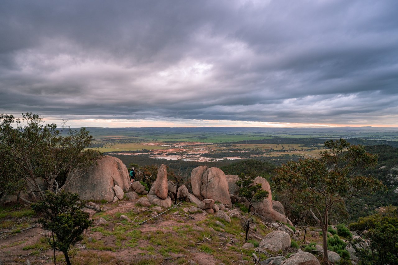 A photo taken from the top of the You Yangs. A cluster of boulders are in the middle of the photo. Beyond them is the view of farmland below the mountain and grey, cloudy skies.