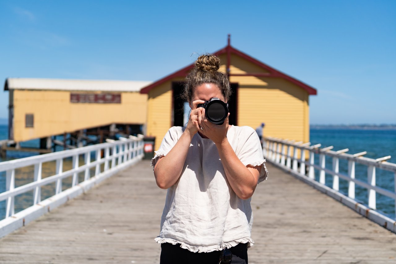 A woman stands at the end of the pier facing the camera. She holds a camera up to her face, posing to take a photo of the photographer. She wears a white t-shirt and her brown hair is in a bun.